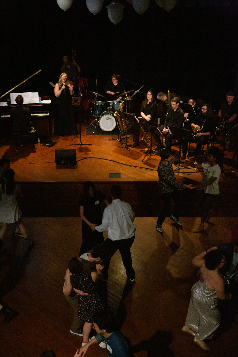 Overhead view of the full band on stage — vocalist, bassist, drummer, and horns — as couples swing dance below.