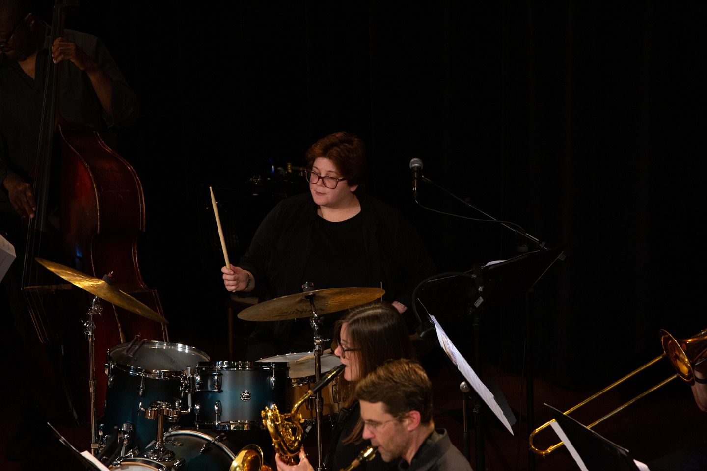 Chelsea Hughey drumming behind the horn section, drumstick raised.