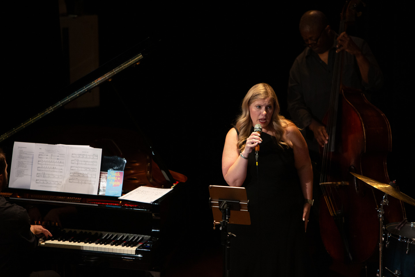 The vocalist singing at the microphone with the grand piano and upright bass behind her.