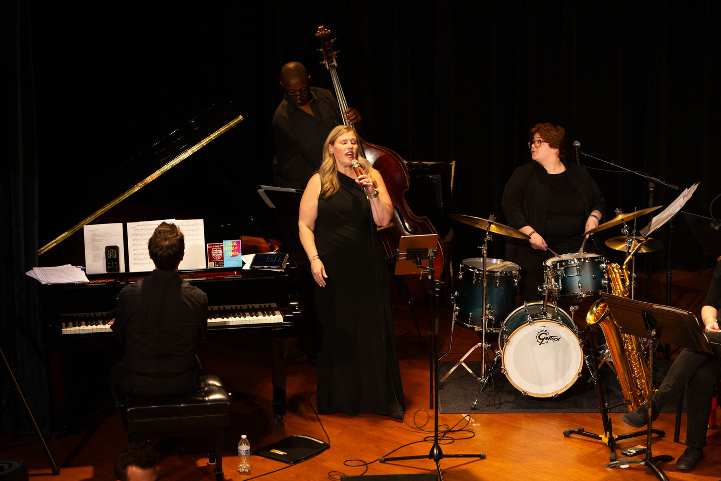 The vocalist singing between the pianist, upright bassist, and drummer on a warmly lit stage.