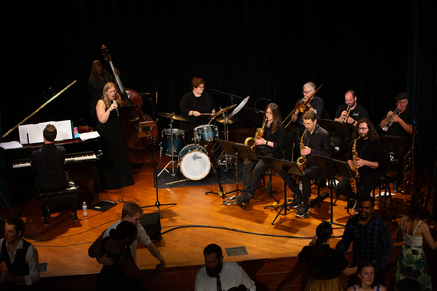 The full 11-piece band on stage — piano, bass, drums, vocals, and horns — as dancers move in the foreground.