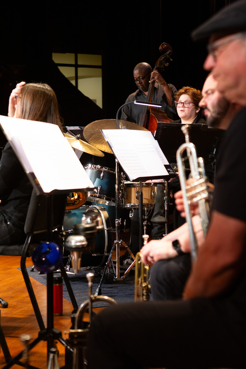 A view from inside the band — a trumpeter in the foreground with drummer and bassist behind the music stands.