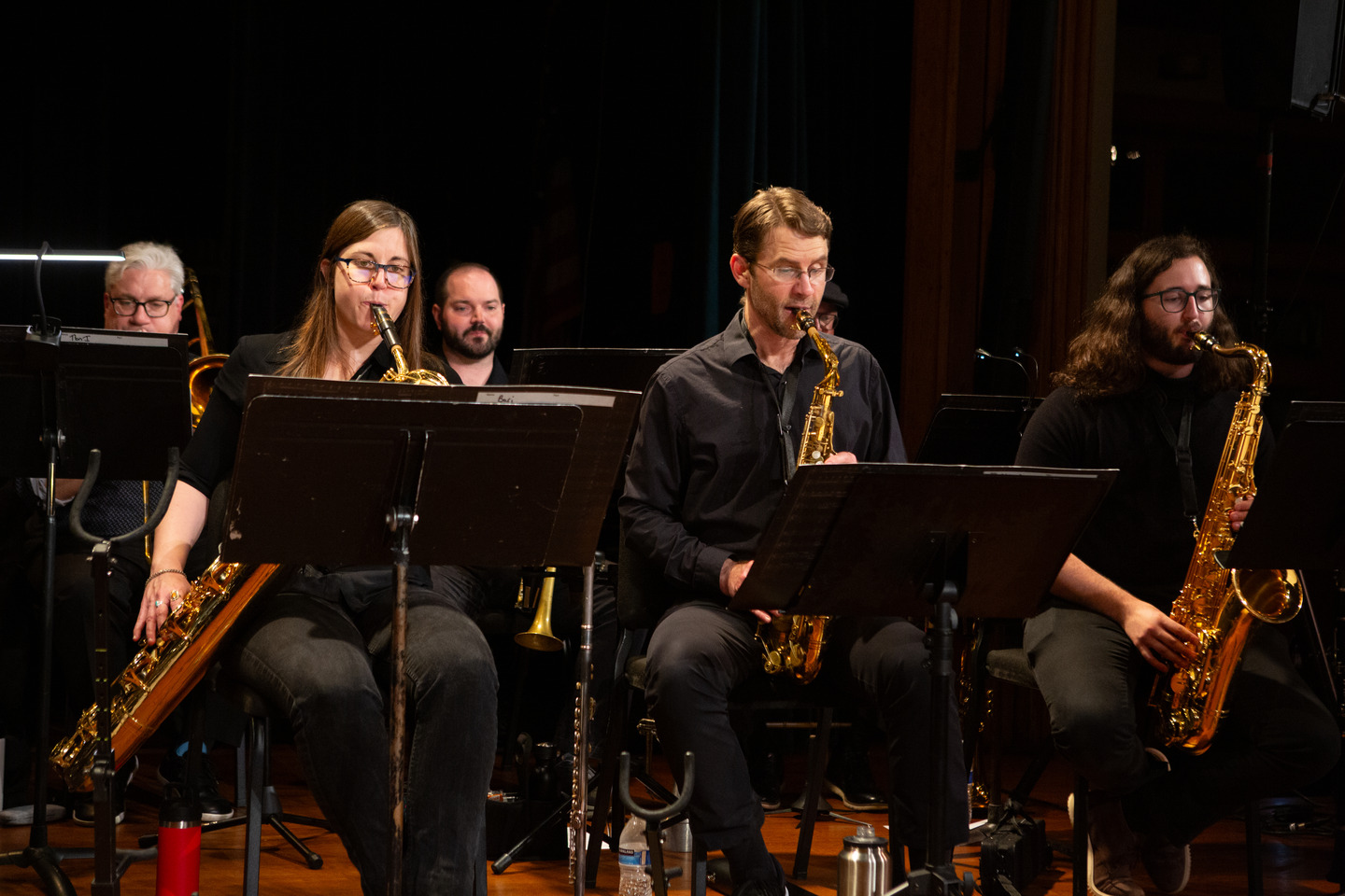 Three saxophonists playing side by side with a trombonist behind them.