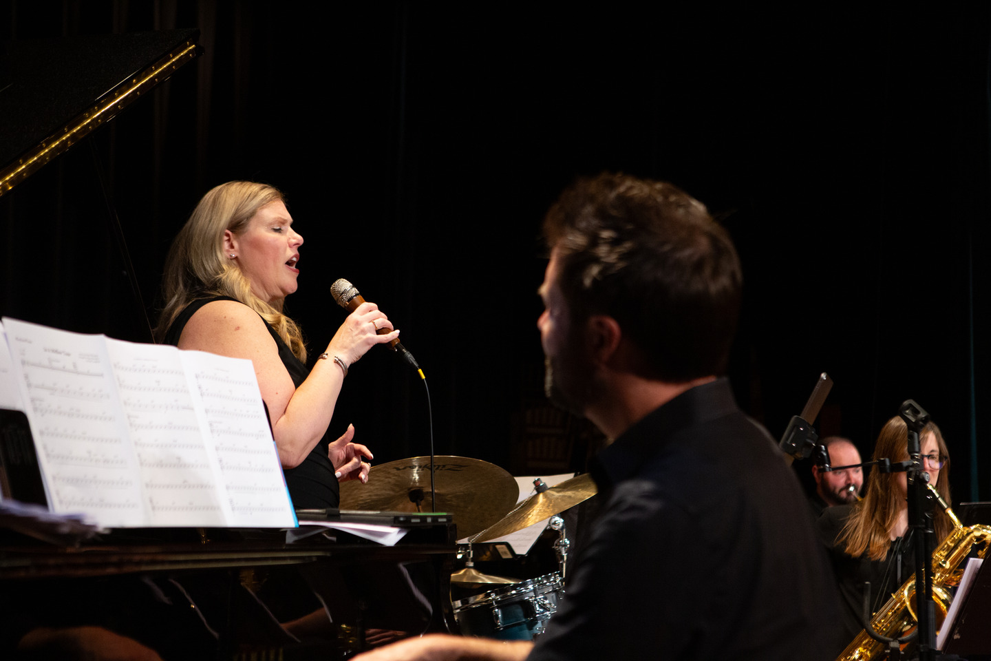 The vocalist singing across the stage toward the pianist, horns visible behind.