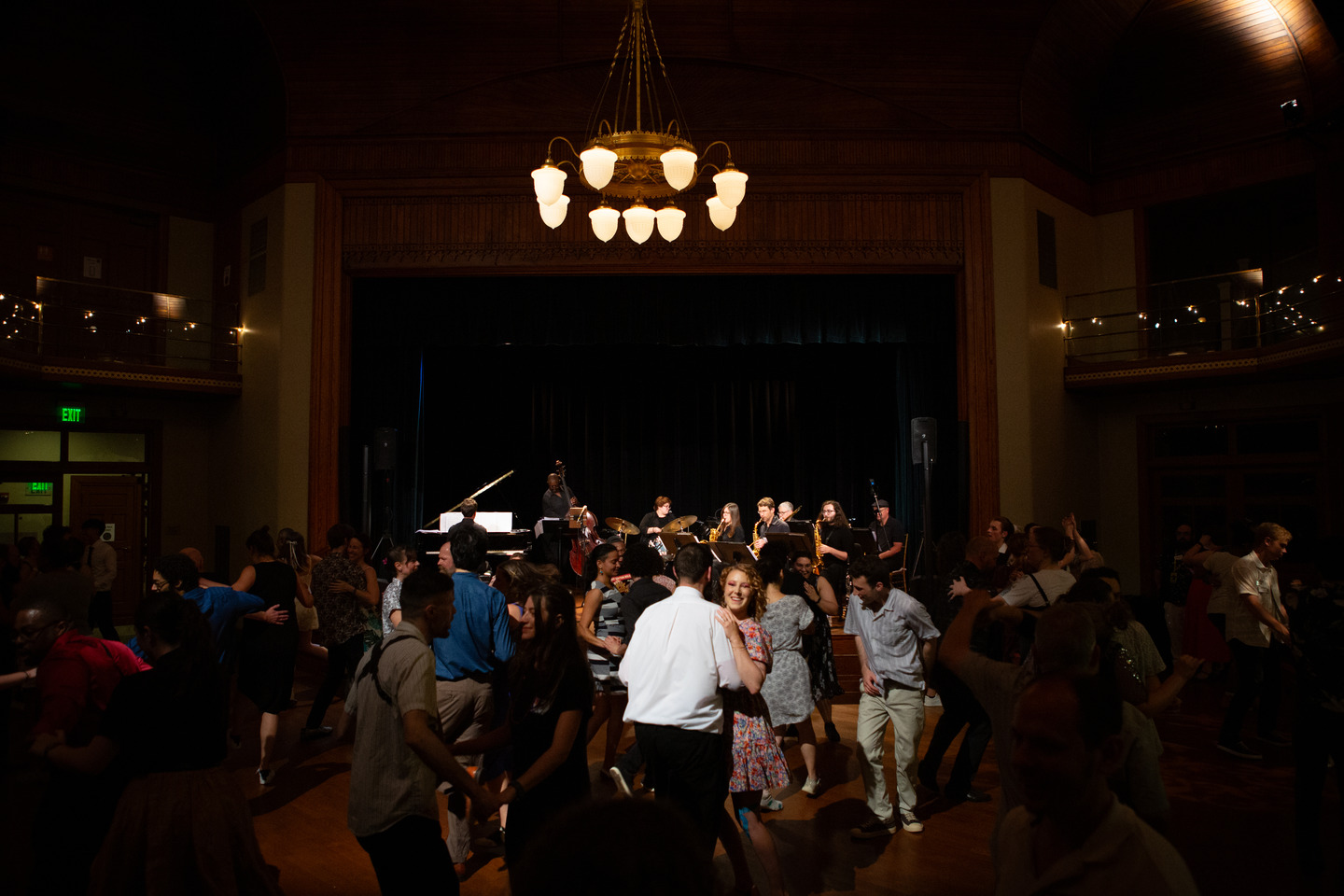 A wide view of the ballroom: the band on the distant stage under a glowing chandelier as the crowd swing dances.
